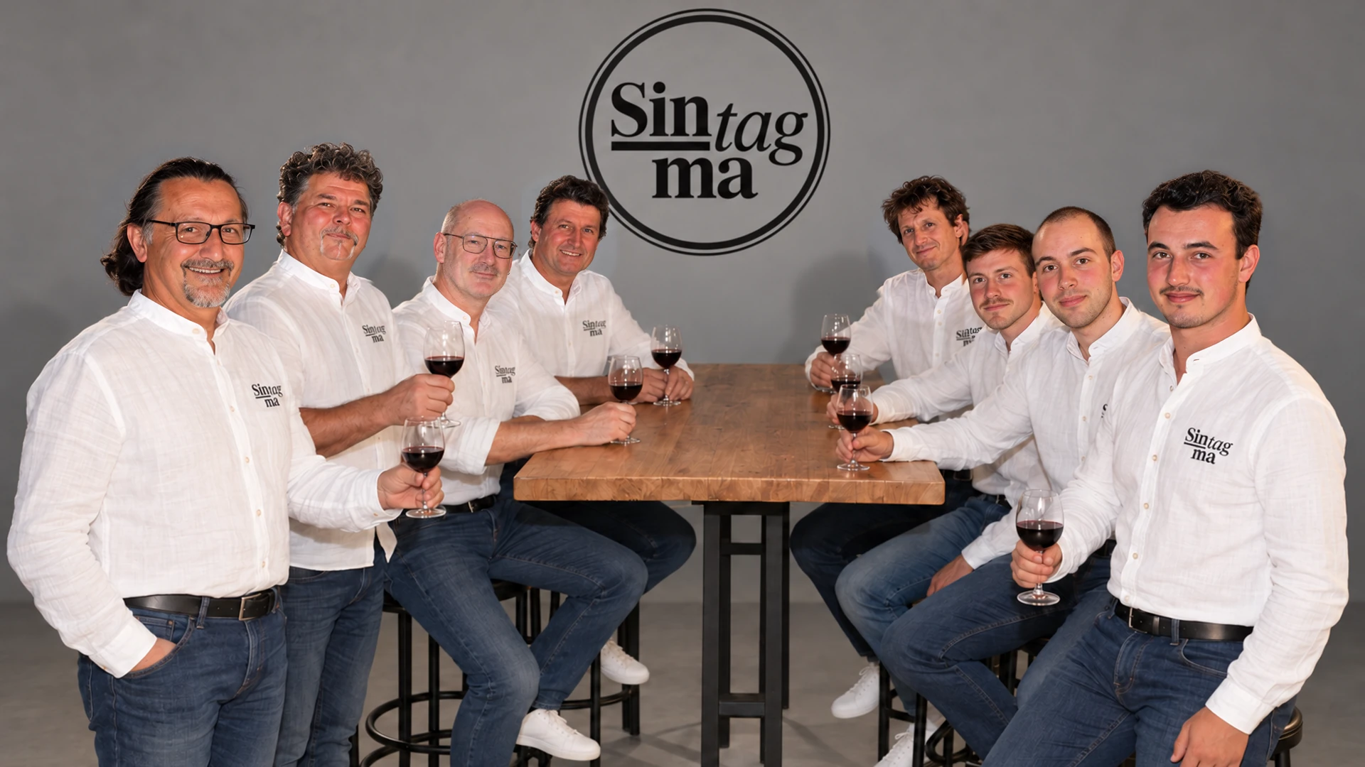 Gli otto Sintagma riuniti al tavolo con un calice di vino, davanti al logo del gruppo